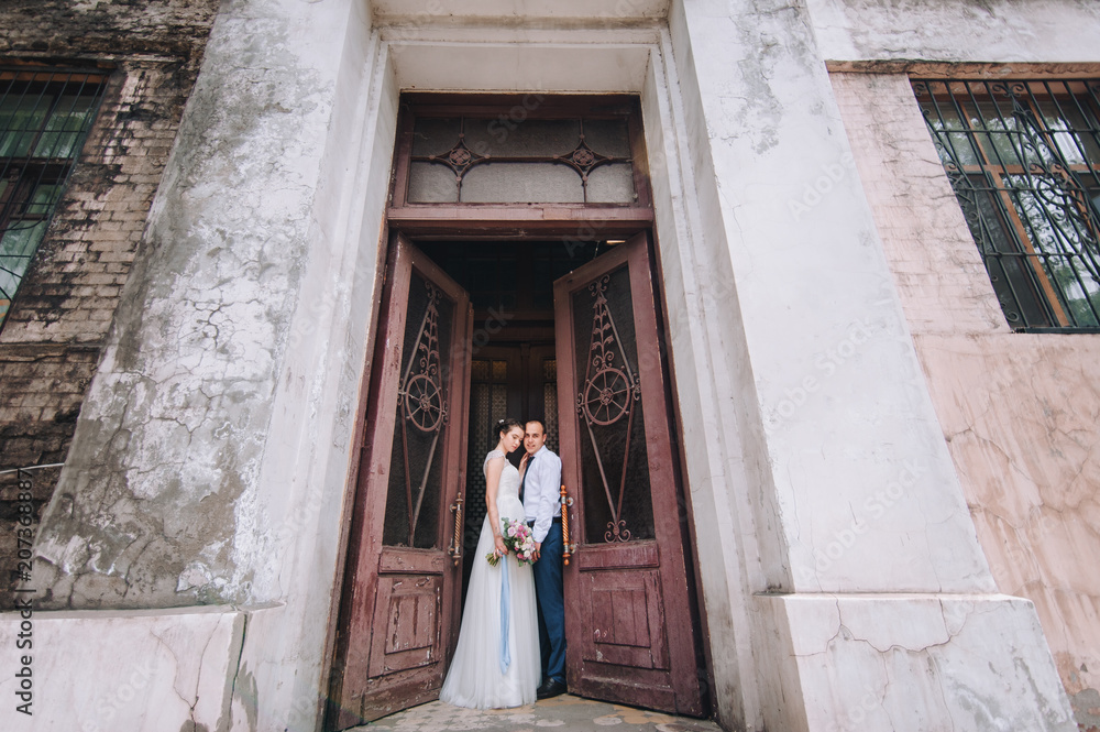 Beautiful newlyweds hug and look at each other in the doorway near an abandoned wall. The stylish bridegroom smiles and holds a sweet bride near the ancient door. Portrait of a young newlywed couple.