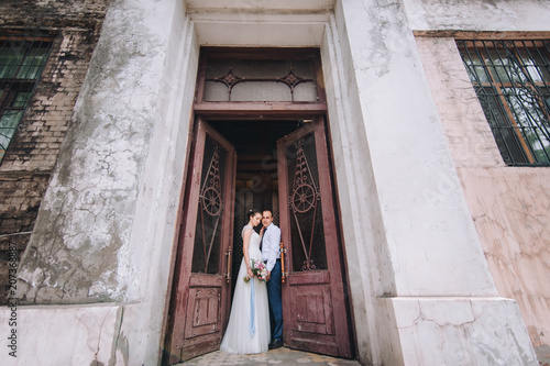 Beautiful newlyweds hug and look at each other in the doorway near an abandoned wall. The stylish bridegroom smiles and holds a sweet bride near the ancient door. Portrait of a young newlywed couple. © shchus