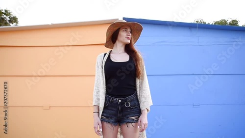 Slow motion of young attractive woman wearing stylish clothes, shorts and straw hat standing and looking at the camera on blue and yellow background. Travel concept. Local souvenir market.