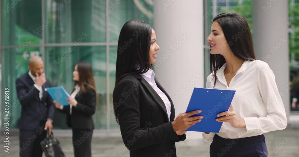 Two business women of different ethnicities talk about finance looking at the patterns of economic markets and banks, and in the background a group of multi-ethnic business people. Two business women of different ethnicities talk about finance looking at the patterns of economic markets and banks, and in the background a group of multi-ethnic business people.