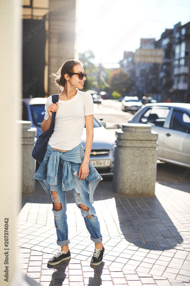 Model wearing plain white t-shirt, boyfriend jeans, sneakers and hipster sunglasses posing against street, teen urban clothing style.