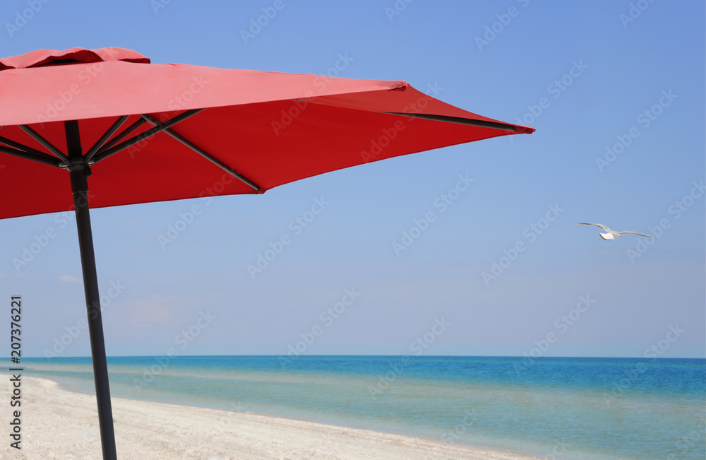 Beach umbrella on a sunny day, sea in background. Beach umbrella close-up. Soaring seagull. Idyllic tropical beach with red umbrella, white sand, turquoise ocean water and blue sky.