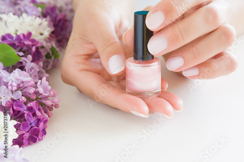 Young, perfect, groomed woman's hands with pink nail varnish bottle. Nails care. Manicure, pedicure beauty salon. Beautiful branches of lilac blossoms on white table. Colorful, fresh flowers.