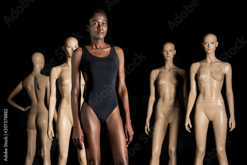 beautiful african american woman in bodysuit standing in front of dummies and looking at camera on black