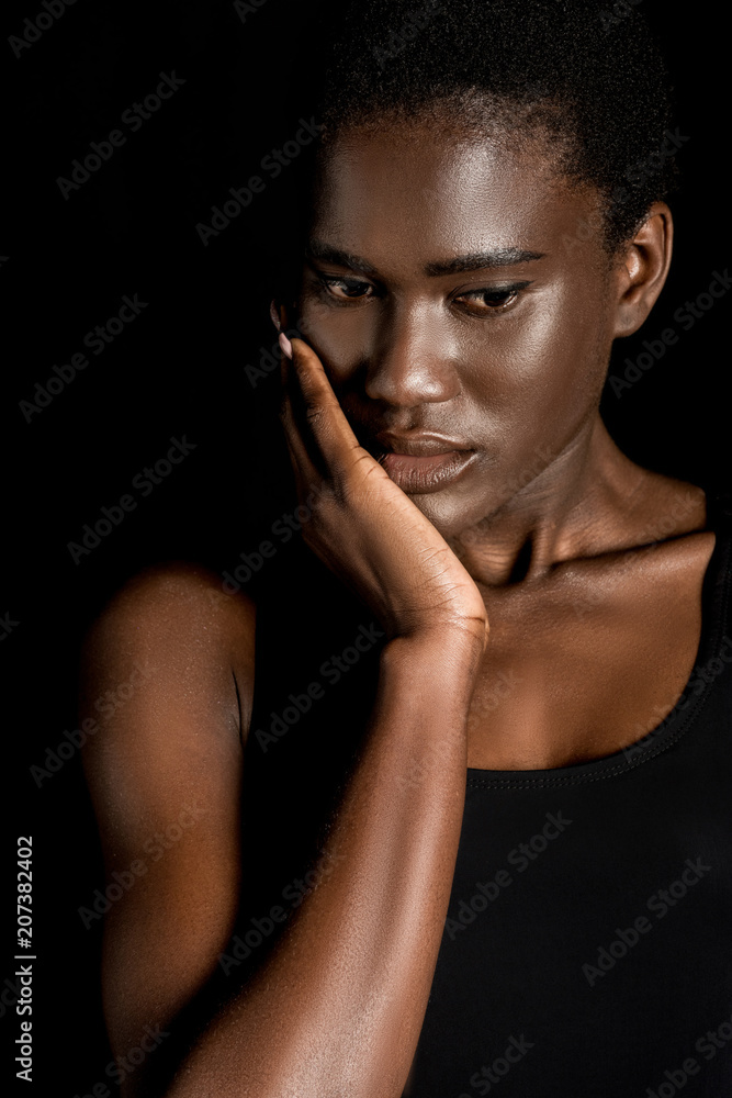 pensive young african american woman touching face with hand and looking down isolated on black