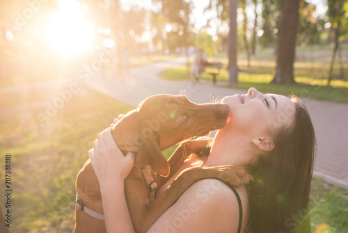 Portrait of a dog kissing a happy girl in a park against the background of the sunset. Leisure with a dog in the park. Girl hugs her with a dog on the summer sunny day at the background of the sun photo