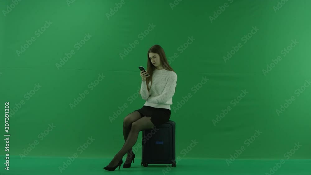 the girl sits in a half-turn on a suitcase , looks at the left, and looks in phone on green screen the girl sits in a half-turn on a suitcase , looks at the left, and looks in phone on green screen
