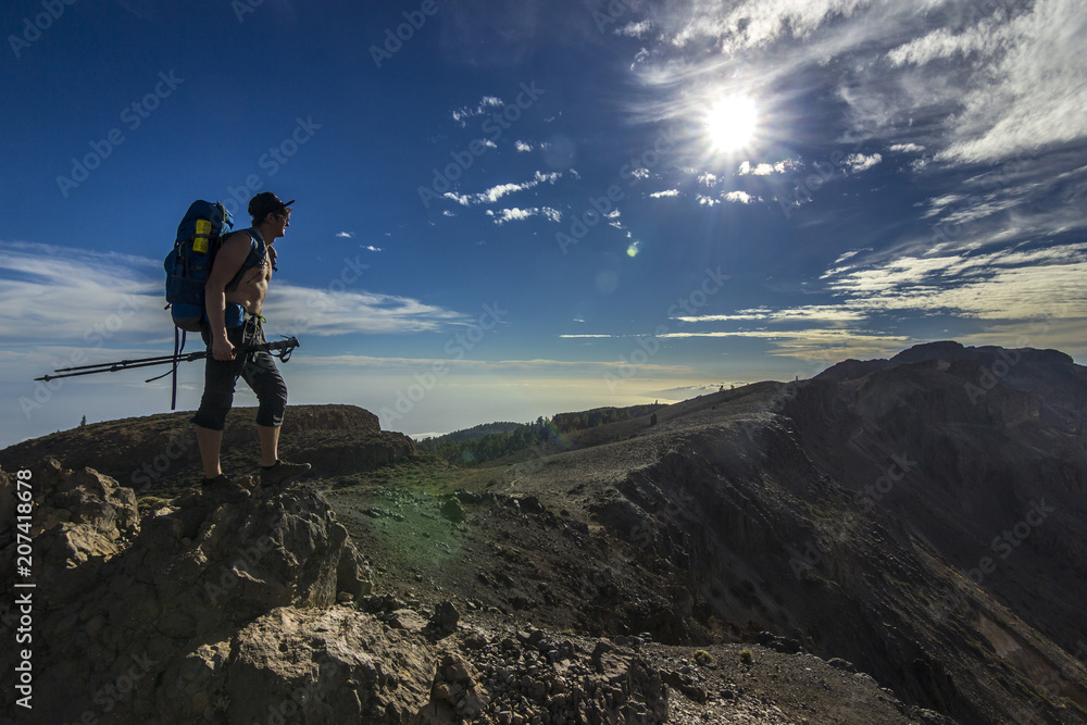 Fototapeta premium man with backpack standing on a cliff in mountains with blue sky and sun