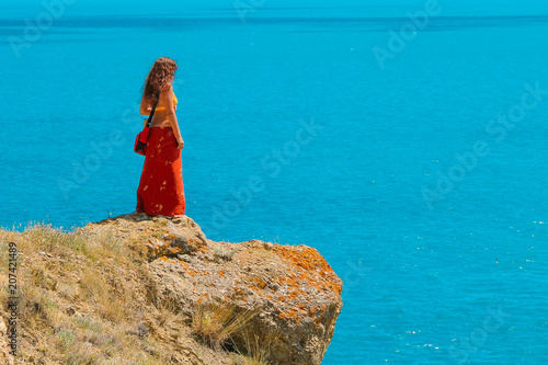 A woman is standing alone and on the edge of a cliff and looking over the blue Sea