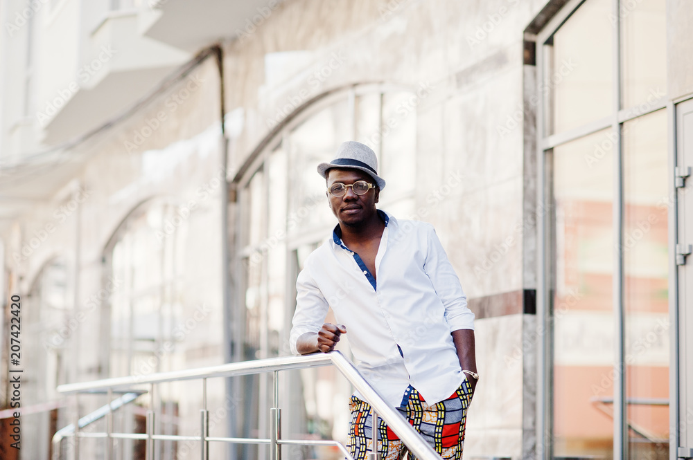 Stylish african american man in white shirt and colored pants with hat and glasses posed outdoor. Black fashionable model boy.