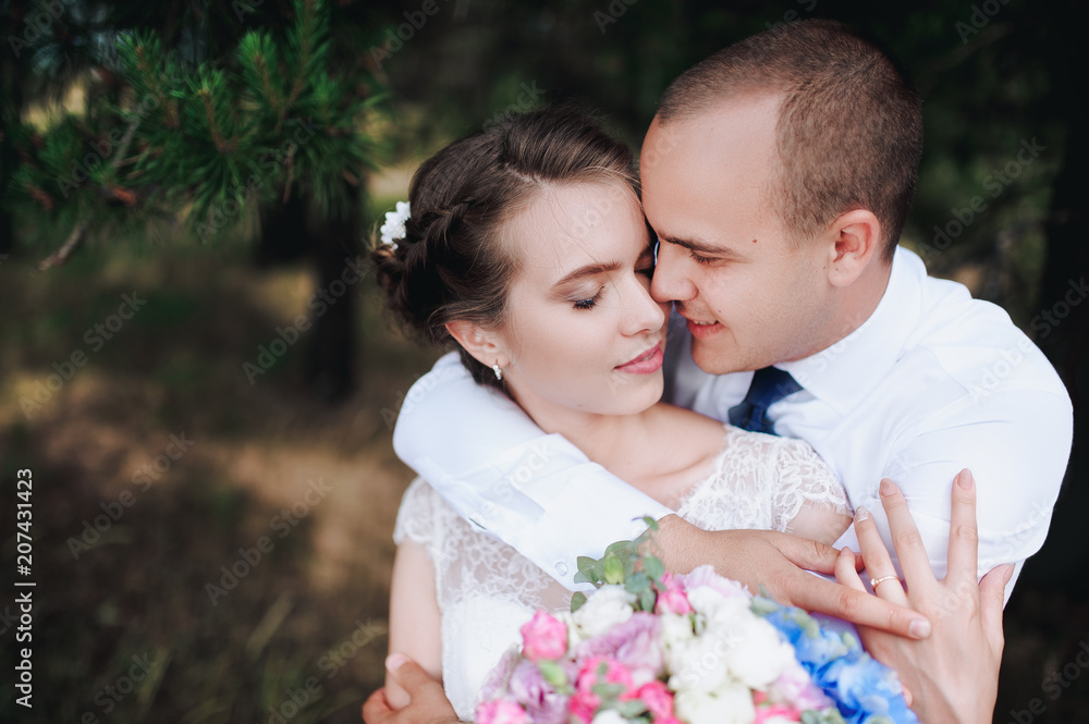 Beautiful and smiling newlyweds gently embrace in a green pine forest on a sunny day. The young groom hugs and kisses from behind a beautiful bride in a lace dress in nature. Wedding portrait close-up Beautiful and smiling newlyweds gently embrace in a green pine forest on a sunny day. The young groom hugs and kisses from behind a beautiful bride in a lace dress in nature. Wedding portrait close-up