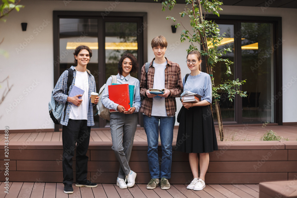Young students standing with books and folders in hands and joyfully looking in camera while spending time together in courtyard of university