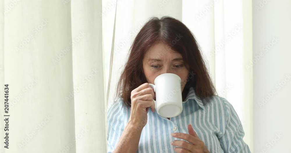 Senior woman resting at home standing near the window drinking tea elderly lifestyle. Happy elderly woman looking at camera Senior woman resting at home standing near the window drinking tea elderly lifestyle. Happy elderly woman looking at camera