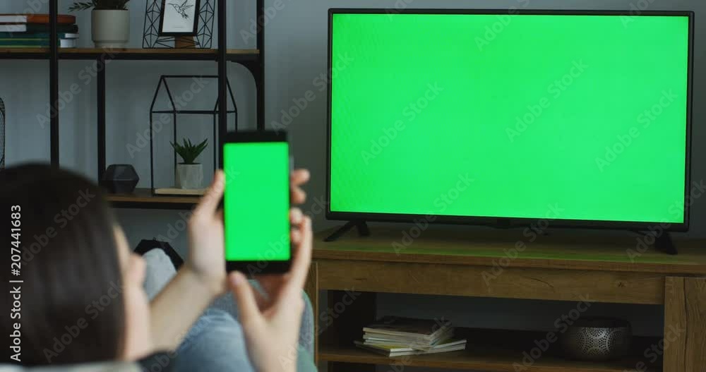 View from the back on the brunette woman resting in the living room, watching TV with green screen and something on the green screen of the smartphone. TV in focus. Chroma key. Indoor View from the back on the brunette woman resting in the living room, watching TV with green screen and something on the green screen of the smartphone. TV in focus. Chroma key. Indoor
