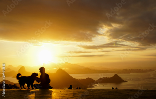Silhueta de um homem sentado fazendo carinho e um cão durante entardecer com montanhas ao fundo.