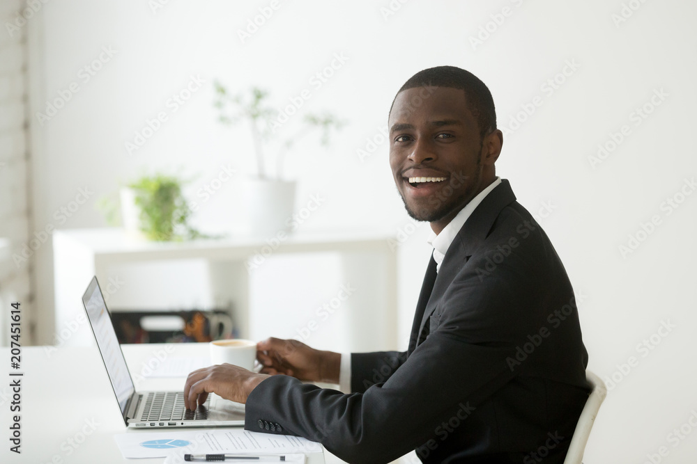 Smiling positive African American looking at camera working at laptop and drinking coffee. Black company financial manager preparing report using computer. Friendly employee posing in small office