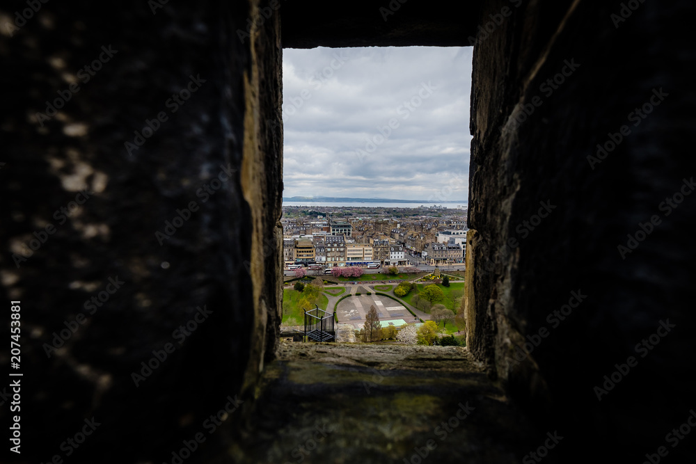 View of Princes Street garden and shops seen through stone windows in Edinburgh Castle during a cloudy day.