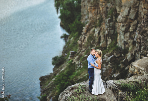 A nice bridegroom embraces a beautiful bride with curly hair and rests her forehead, standing on a precipice. Lovely newlyweds stand on a rock, against the background of rocks and the sea.