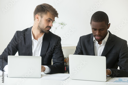 Curious Caucasian male businessman looking at screen of African American colleague busy working at laptop, worker glancing at rival computer, being smart, sly, cunning. Concept of rivalry cooperation photo