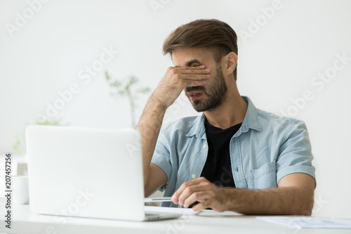 Exhausted casual sick male worker massaging eyes, suffering from eyes strain working long hours in front of laptop screen, relieving tension. Concept of overwork, dizziness, blurry vision