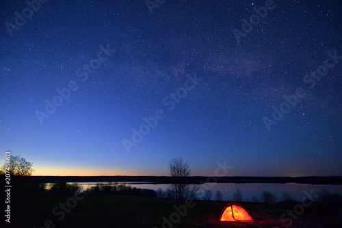 Night landscape with starry sky, Milky Way, lake in the background, and red illuminated tent in the foreground.