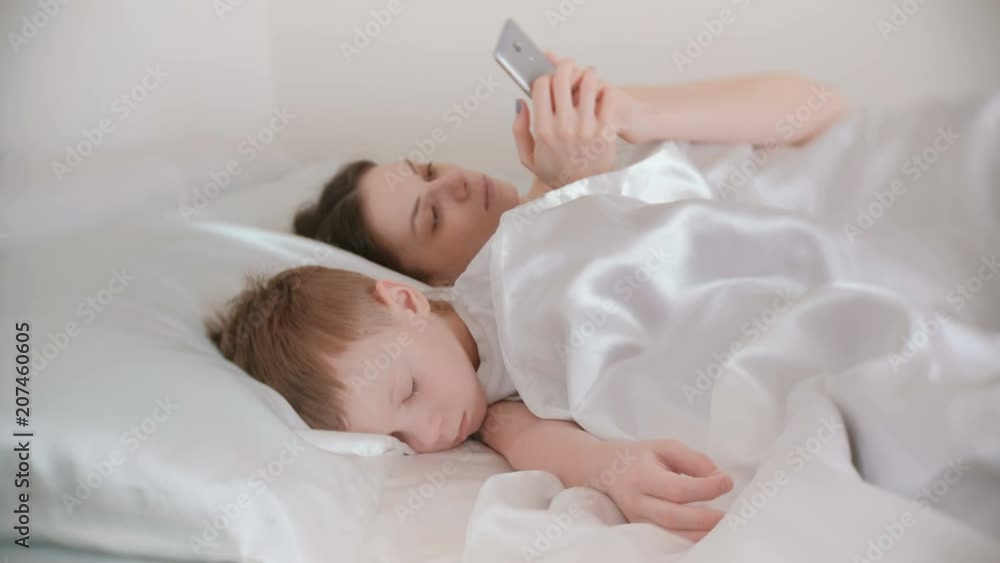 Mom woke up and browsing internet in her mobile phone while her son is still asleep. Mom woke up and browsing internet in her mobile phone while her son is still asleep.