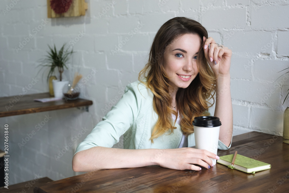 Beautiful young girl sitting at the table with coffee in hand and smiling looking at the camera. Horizontal photo Beautiful young girl sitting at the table with coffee in hand and smiling looking at the camera. Horizontal photo