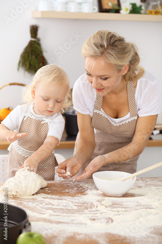 Little girl and her blonde mom in beige aprons playing and laughing while kneading the dough in the kitchen. Homemade pastry for bread, pizza or bake cookies