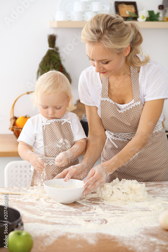 Little girl and her blonde mom in beige aprons  playing and laughing while kneading the dough in the kitchen. Homemade pastry for bread, pizza or bake cookies