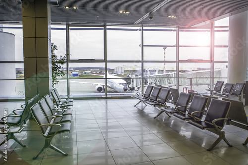 Airplane  view from airport terminal. Modern airport terminal with black leather seats on a sunny morning. A huge viewing glass facade with a passenger aircraft behind it.