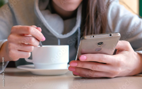 Woman is drinking a hot chocolate with spoon and look on the screen of mobile phone sitting in cafe. Closeup woman's hands.