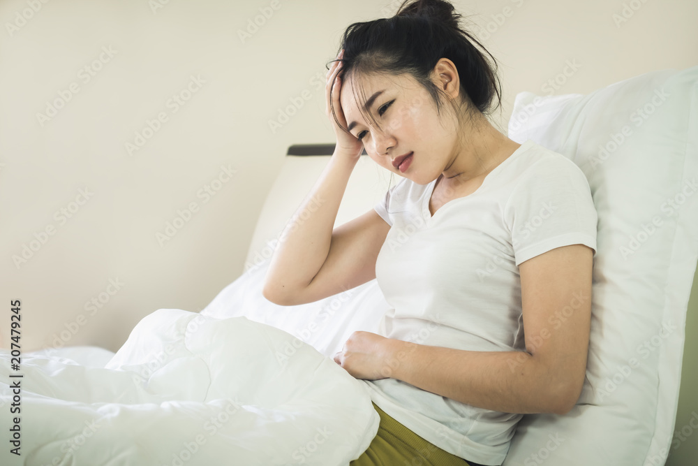 Asian young woman suffering from headache and abdominal pain while sitting on bed at home Asian young woman suffering from headache and abdominal pain while sitting on bed at home