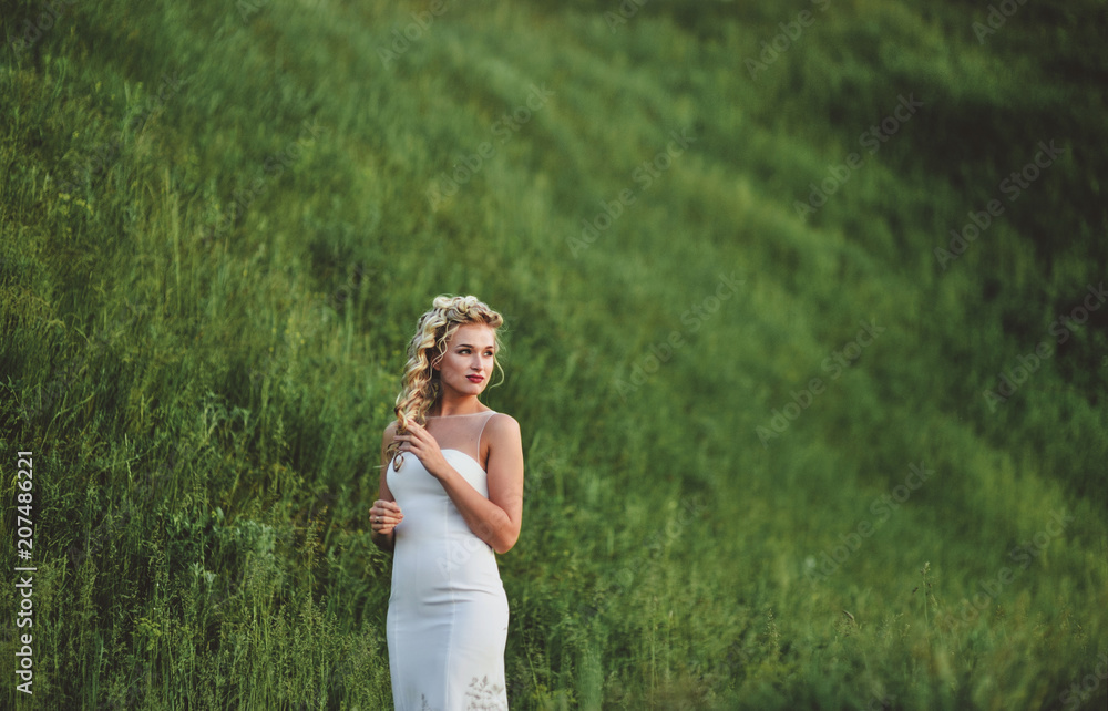 A beautiful and young bride with curly hair is standing in the green grass and enjoying her hairdo. Portrait of a gentle bride. Evening wedding. Model at the wedding.