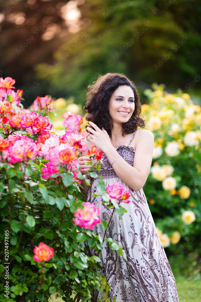 A vertical portrait of young Caucasian woman with dark brown curly hair near pink rose bushes, looking to her left, smiling with teeth, in summer dress A vertical portrait of young Caucasian woman with dark brown curly hair near pink rose bushes, looking to her left, smiling with teeth, in summer dress