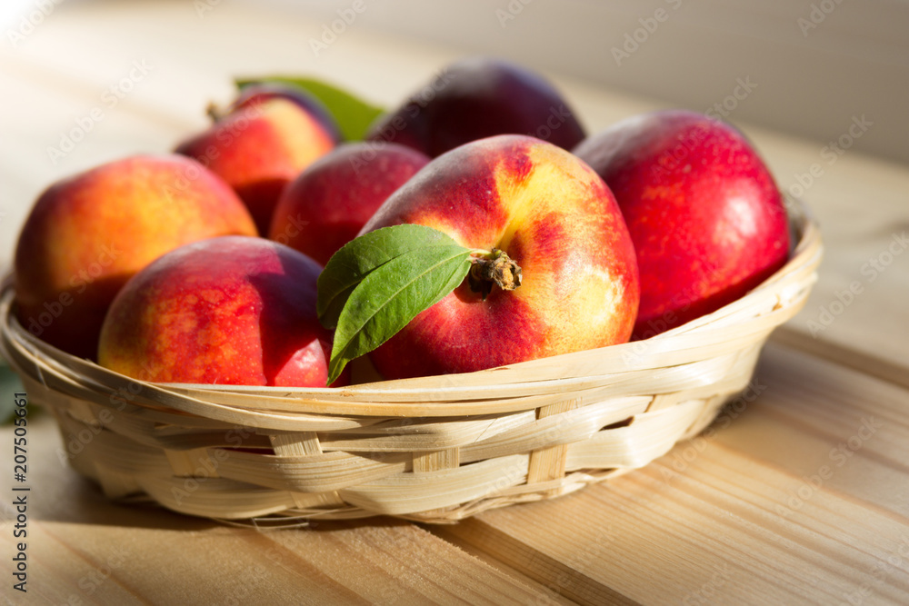 fresh nectarine with leaves  are in a wicker plate on a light wooden table in the sun. close-up