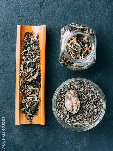 Dry green tea leaves on a wooden spoon and in a glass jar on a dark background.