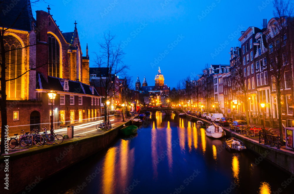 Naklejka premium Historical old residential house building along river water canal of amsterdam during sunrise with lights and empty street pavement under cloudy warm sky with church background