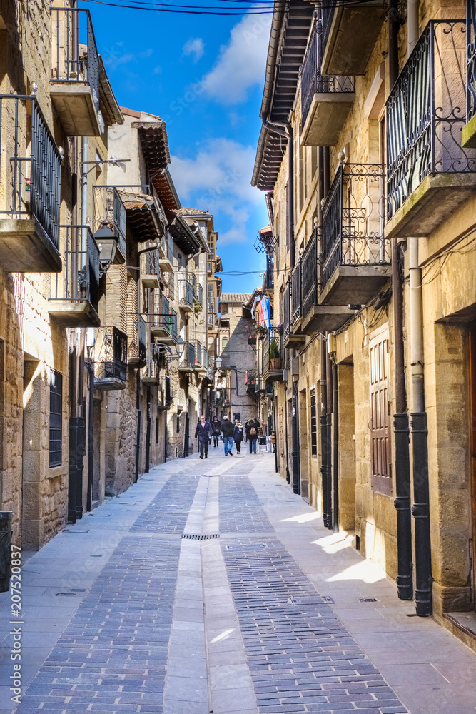  Main street of the town called Mayor with facades of stone houses and iron balconies typical of northern Spain and people passing through it. Stone paving floor