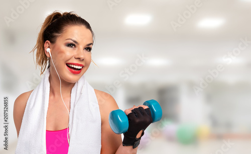 Fitness activity banner - beautiful young woman in sportswear with towel, earphones and dumbbells making workout on a blurred background of a gym (copy space)