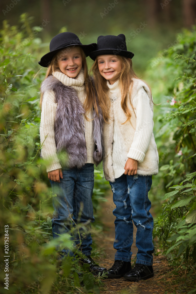 Two happy girls are equally dressed: in fur vests and hats in the forest. Little girlfriends in park. Children Friendship Together Smiling Happiness Concept