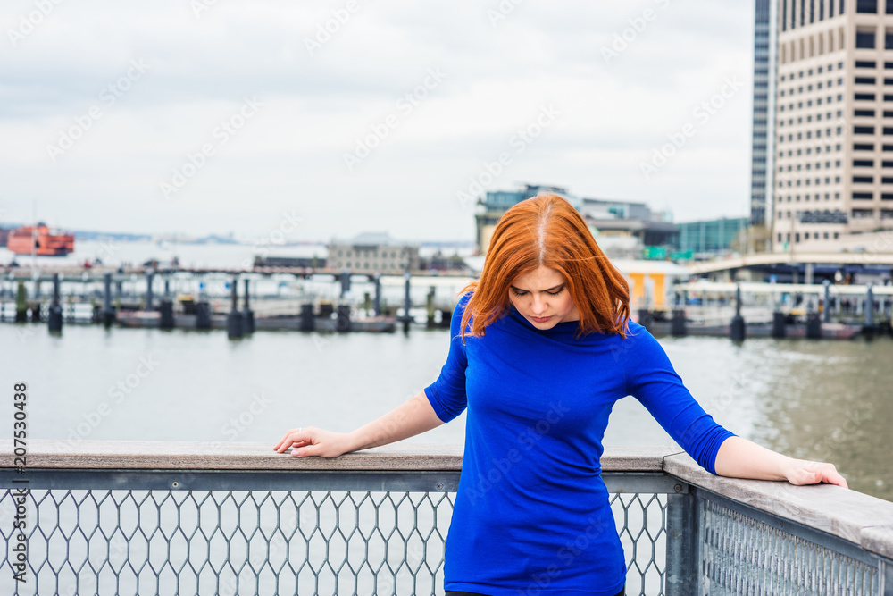 Young American Woman traveling in Manhattan, New York, wearing blue long sleeve T shirt, standing by river at harbor in business district under cloudy sky, lowering head, sad, thinking..