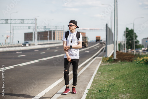 Stylish man hitchhiker on the street or motorway. He wearing in black jeans, black T-shirt, black cap with round sunglasses © Northern life