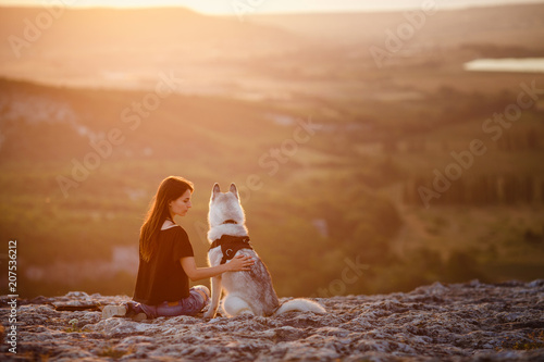 Beautiful girl plays with a dog, grey and white husky, in the mountains at sunset