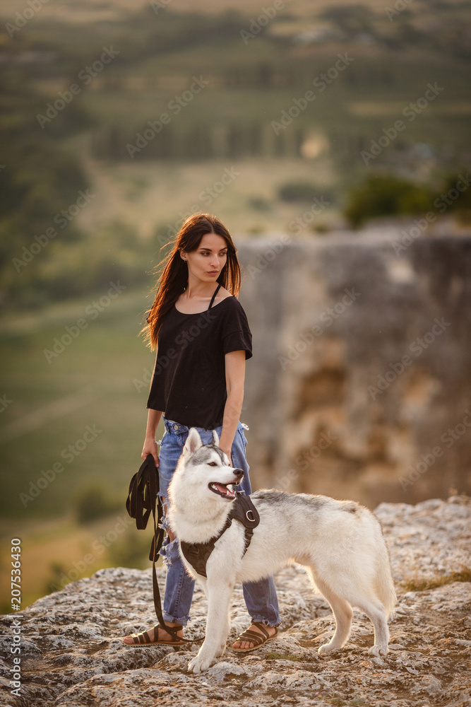 Beautiful girl plays with a dog, grey and white husky, in the mountains at sunset.
