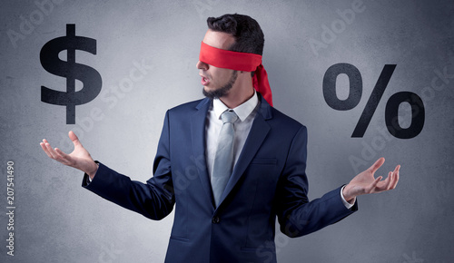 Serious businessmen standing in front of a grey wall with red ribbon on his eye, holding dollars 