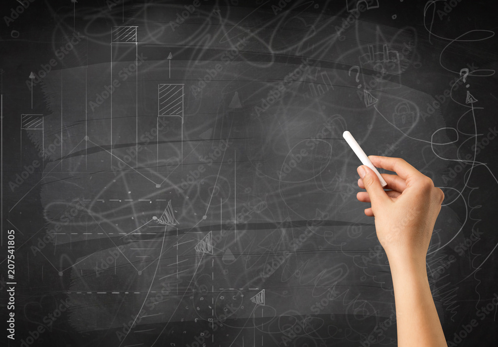 Fototapeta premium Female hand holding white chalk in front of a blackboard with scribbles and plans drawn on it 