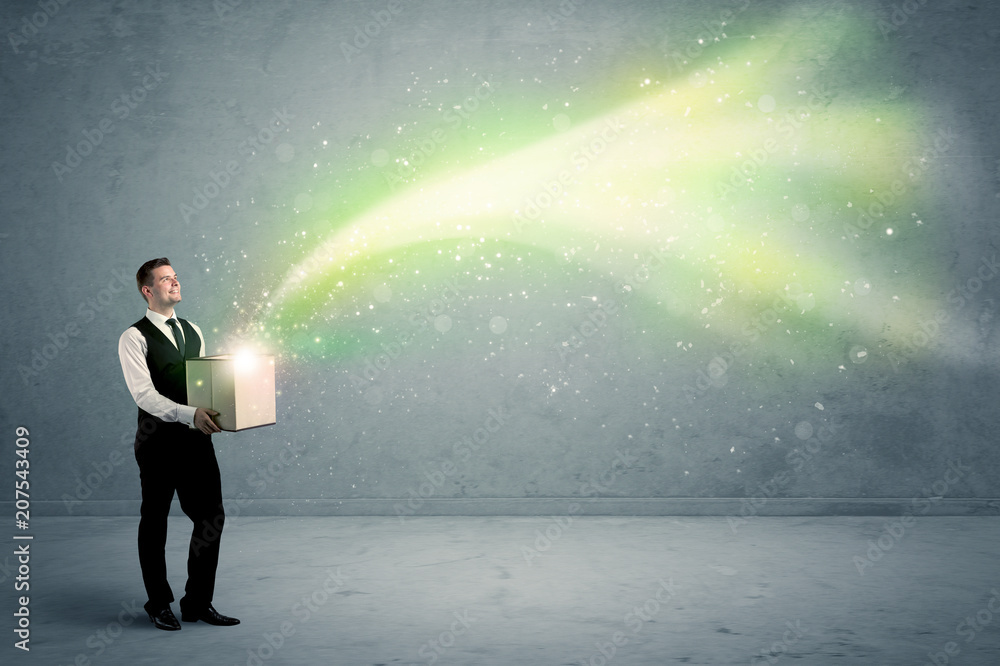 Bright yellow, green light beams escaping a cardboard box held by young elegant male business person in stylish suit concept. Bright yellow, green light beams escaping a cardboard box held by young elegant male business person in stylish suit concept.