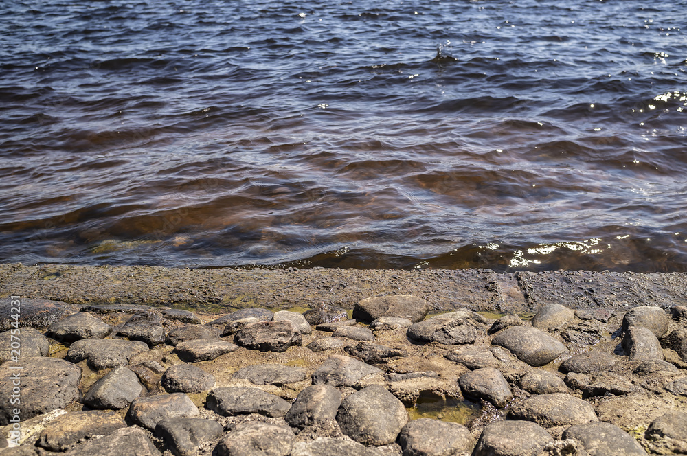 Fototapeta premium An ancient embankment of cobblestone in the background of a river with clear water and waves in the rays of the spring sun.