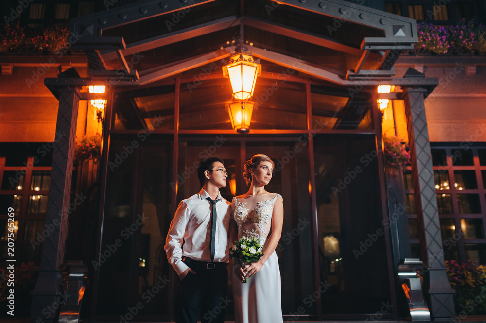 Stylish newlyweds on the background of the hotel. Night wedding walk. International wedding of Asian and European.