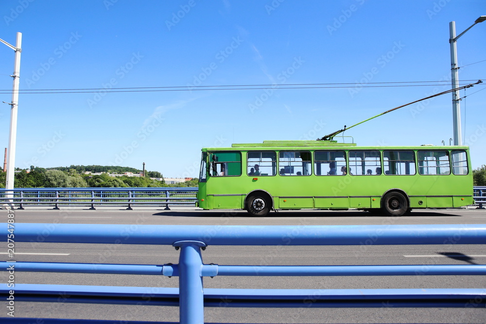 Fototapeta premium Trolley bus crossing bridge. Green trolleybus moving on road against blue barrier or guard rail.